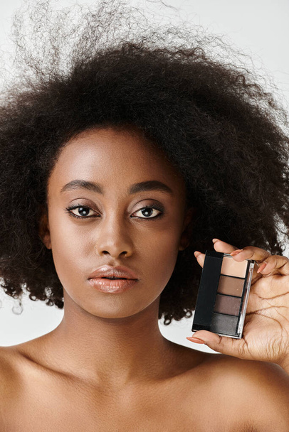 Μια νεαρή Αφρο-Αμερικανίδα με σγουρά μαλλιά που κρατάει μια παλέτα μακιγιάζ σε ένα στούντιο, δίνοντας έμφαση στη φροντίδα του δέρματος. - Φωτογραφία, εικόνα