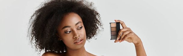 Μια όμορφη νεαρή Αφροαμερικανή με σγουρά μαλλιά σε ένα στούντιο, κρατώντας παλέτα μακιγιάζ - Φωτογραφία, εικόνα