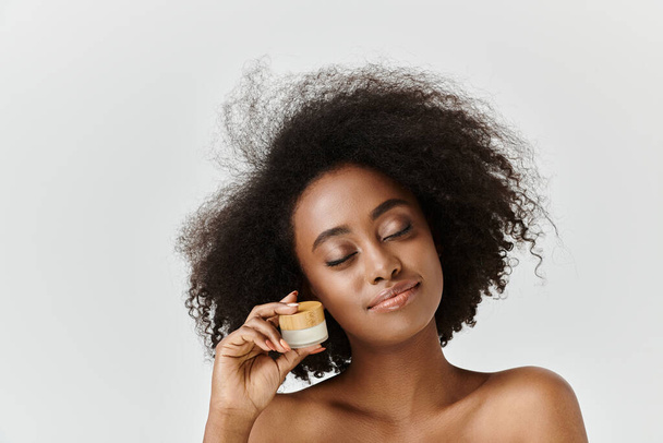 Μια όμορφη νεαρή Αφρο-Αμερικανίδα με σγουρά μαλλιά κρατώντας ένα βάζο με κρέμα στο δεξί της χέρι.. - Φωτογραφία, εικόνα
