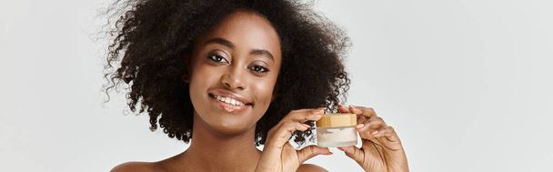 Una bella giovane donna afroamericana con i capelli ricci che tiene in mano un barattolo di crema, promuovendo la cura della pelle. - Foto, immagini