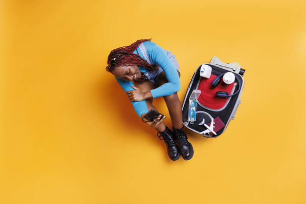 アフリカ系アメリカ人の少女は,スタジオの床に座っている携帯電話を使用し,彼女の隣の旅行の必需品でいっぱいの大きなトロリーバッグを持っています. 離れる準備をしているスマートフォンを持つ若い大人. - 写真・画像