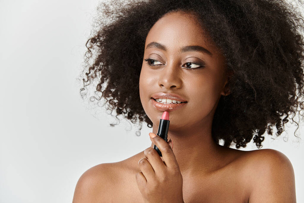 Una hermosa joven afroamericana con el pelo rizado sosteniendo un lápiz labial en la mano, centrándose en la belleza. - Foto, Imagen