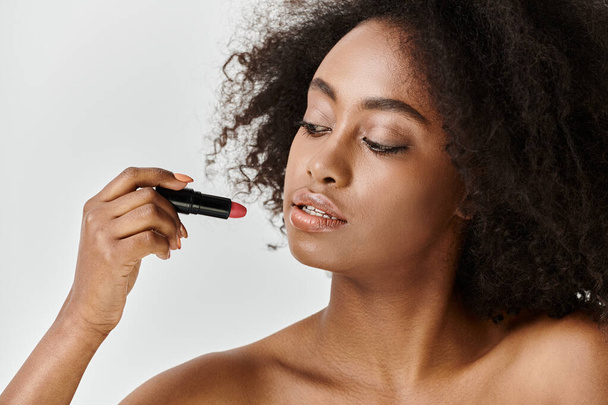 Una joven afroamericana con el pelo rizado se está aplicando hábilmente lápiz labial en la cara en un ambiente de estudio. - Foto, Imagen
