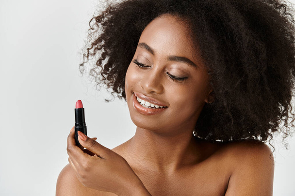 Piękna młoda Afroamerykanka z kręconymi włosami trzyma szminkę w dłoni, ucieleśniając koncepcję pielęgnacji skóry. - Zdjęcie, obraz