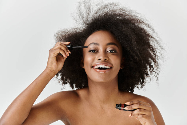 Une belle jeune femme afro-américaine avec ses cheveux bouclés dans un décor de studio, appliquant le mascara - Photo, image
