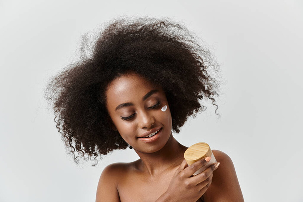 Eine elegante Afroamerikanerin mit voluminöser Afro-Frisur hält in einem Studio anmutig eine Creme in der Hand. - Foto, Bild