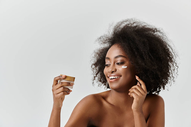 Una joven afroamericana con el pelo rizado sonríe mientras sostiene un frasco de crema, irradiando alegría y satisfacción. - Foto, imagen