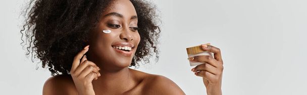 Μια όμορφη νεαρή Αφροαμερικανή γυναίκα με σγουρά μαλλιά απλώνει κρέμα στο πρόσωπό της, εστιάζοντας στη φροντίδα του δέρματος. - Φωτογραφία, εικόνα