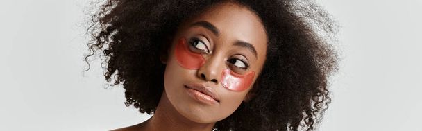 Ohromující mladá Afroameričanka s kudrnatými vlasy ztělesňuje koncepci péče o pleť a zároveň má nápadnou červenou pásku přes oko. - Fotografie, Obrázek