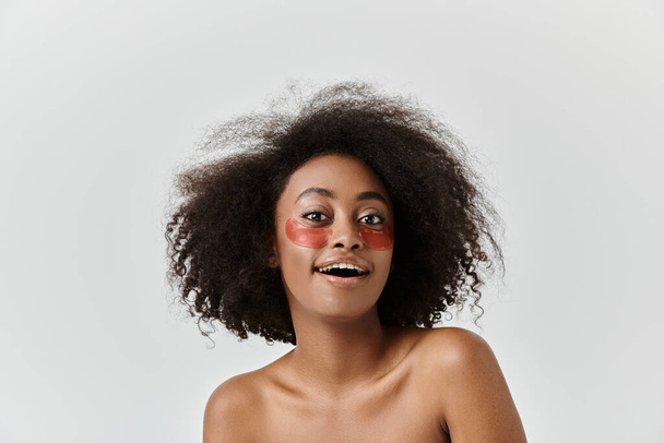 Μια όμορφη νεαρή Αφροαμερικανή γυναίκα με σγουρά μαλλιά που επιδεικνύει κάτω από τα μπαλώματα των ματιών, ενσαρκώνοντας δημιουργικότητα και αυτοέκφραση. - Φωτογραφία, εικόνα