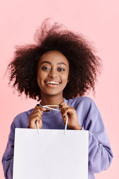 A beautiful young African American woman with curly hair happily holds a shopping bag in a studio setting. - Photo, Image