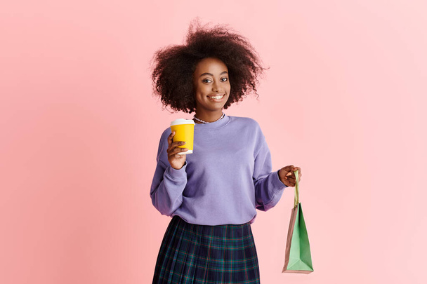 Стильная афроамериканка с вьющимися волосами держит чашку кофе и бумажный пакет в модной обстановке студии. - Фото, изображение