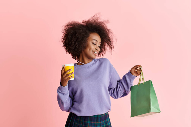 Egy fiatal afro-amerikai nő göndör hajjal, kezében egy csésze kávéval és egy bevásárlószatyorral egy stúdióban.. - Fotó, kép