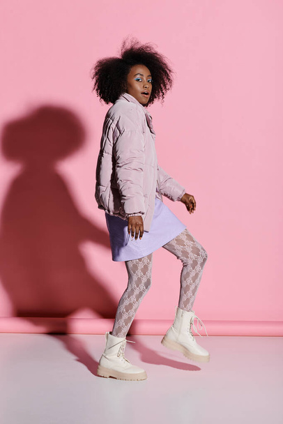Стильная молодая афроамериканка с вьющимися волосами в розовой куртке и белой юбкой наносит позу перед розовой стеной.. - Фото, изображение