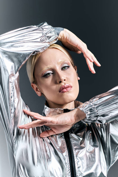appealing woman with blonde hair in futuristic silver attire posing in motion on gray backdrop - Фото, изображение