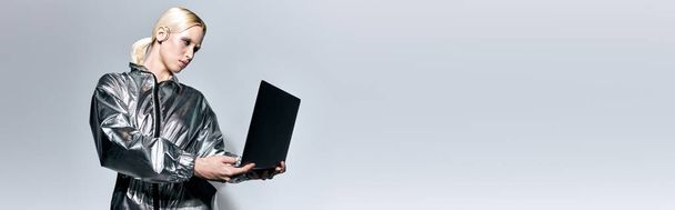 extraordinaria mujer con maquillaje en ropa futurista mirando a su computadora portátil en fondo gris, pancarta - Foto, Imagen
