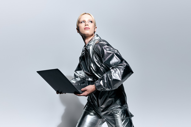 extravagant beautiful woman in silver robotic clothing posing in motion with laptop and looking away - Foto, afbeelding