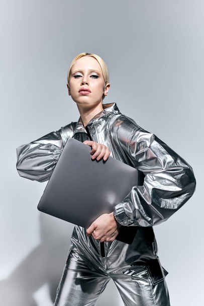beautiful blonde female model in robotic silver outfit holding laptop and looking at camera - Foto, afbeelding