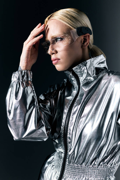 attractive blonde woman with sci fi glasses in futuristic attire looking away on dark gray backdrop - 写真・画像