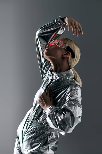 extravagant woman with futuristic glasses in silver attire doing robotic movements and looking away - Foto, imagen