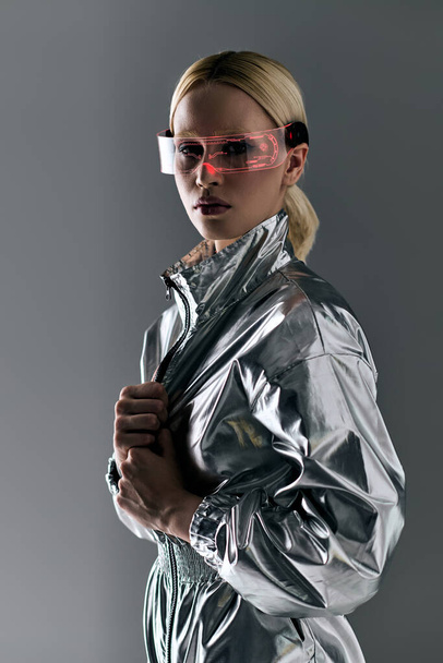 appealing blonde woman with futuristic glasses in silver outfit looking at camera on gray background - Фото, изображение
