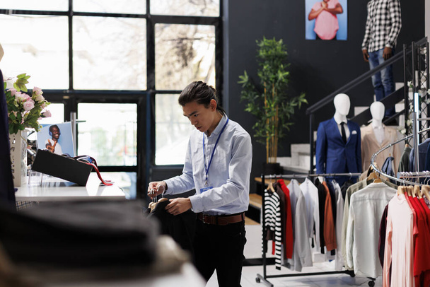 Asyalı mağaza çalışanı kıyafetleri düzenliyor, askılara moda ürünler takıyor, modern butik mağazasında çalışıyor. Alışveriş merkezinde şık bir adam resmi giysileri kontrol ediyor. Moda konsepti - Fotoğraf, Görsel