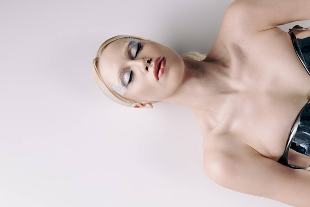 attraktive bizarre Frau mit blonden Haaren und Make-up in silberner futuristischer Kleidung auf dem Boden liegend - Foto, Bild