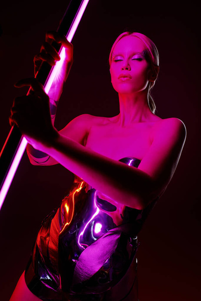 good looking peculiar female model in metallic futuristic attire holding vibrant pink LED lamp stick - Photo, Image