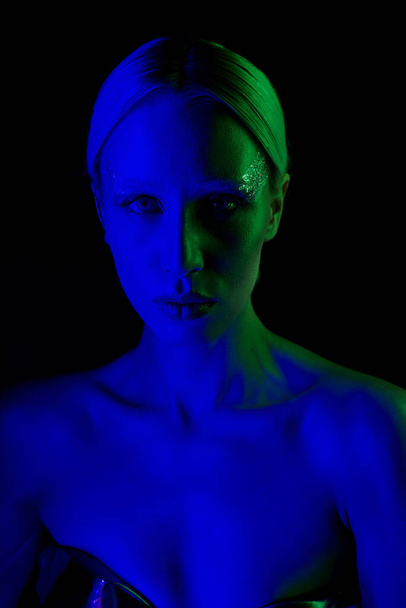 beautiful futuristic woman in metallic sci fi clothing posing in blue lights and looking at camera - Foto, afbeelding