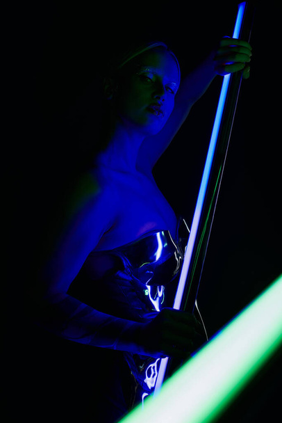atraktivní bizarní žena v robotické oblečení pózování s modrým LED lampa tyč a při pohledu na kameru - Fotografie, Obrázek