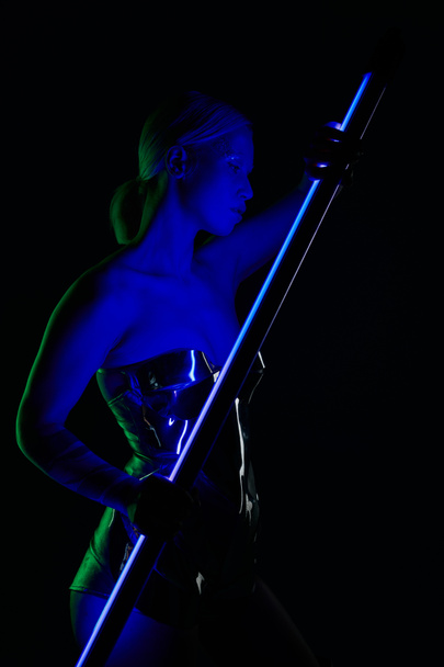 beautiful peculiar woman in silver robotic attire posing with blue LED lamp stick and looking away - Photo, Image