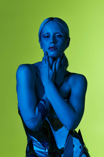 außergewöhnliche Frau in Roboterkleidung posiert in blauem Licht vor grünem Hintergrund und blickt in die Kamera - Foto, Bild