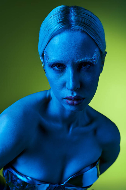 extravagantní žena v robotickém oděvu pózující v modrých světlech na zeleném pozadí a dívající se do kamery - Fotografie, Obrázek