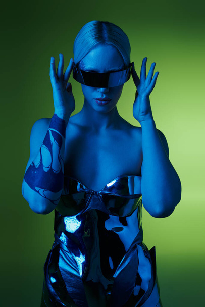 futuristische Frau in silberner Roboterkleidung mit Science-Fiction-Sonnenbrille posiert in Lichtern vor grünem Hintergrund - Foto, Bild