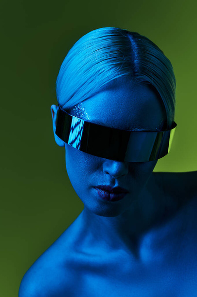 mujer rubia extravagante con gafas de sol robóticas de ciencia ficción posando en luces azules sobre fondo verde - Foto, Imagen