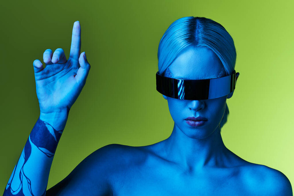 εξαιρετική ξανθιά γυναίκα με sci fi ρομποτικά γυαλιά ηλίου ποζάρουν σε μπλε φώτα σε πράσινο φόντο - Φωτογραφία, εικόνα