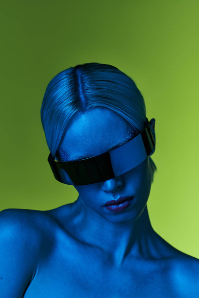 futuristische blonde vrouw met sci fi robotachtige zonnebril poseren in blauw licht op groene achtergrond - Foto, afbeelding