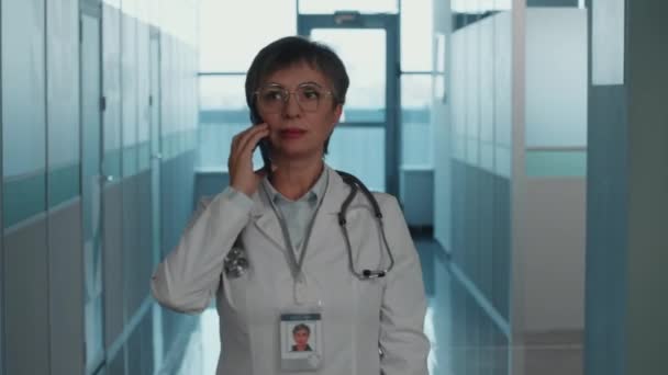 Médico femenino maduro en bata blanca con estetoscopio sobre el cuello e insignia de nombre caminando por el pasillo del hospital, hablando por teléfono móvil y comprobando la hora en el reloj de pulsera - Metraje, vídeo