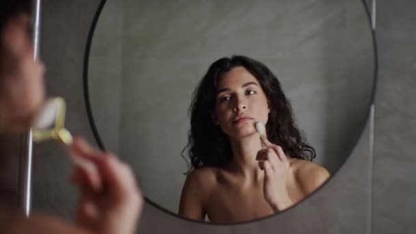Keskipitkä lähikuva yli olkapään laukaus nuori valkoihoinen nainen kihara tukka ja paljain rinnassa seisoo peilin edessä kylpyhuoneessa ja tekee kasvojen hieronta gua sha rulla - Materiaali, video