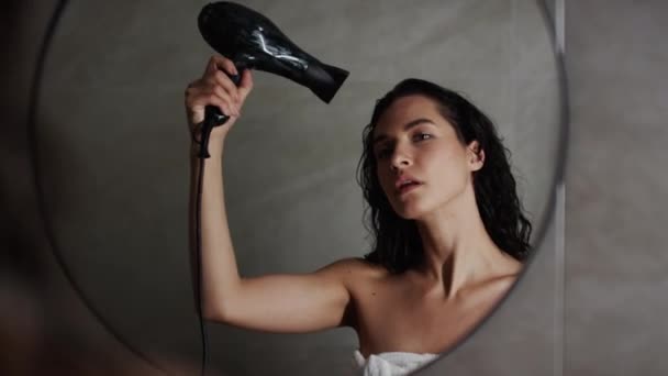 Keskipitkä lähikuva olkapään yli kuva nuoresta kiharasta valkoihoisesta tai latinalaisamerikkalaisesta naisesta, joka seisoo peilin edessä kylpyhuoneessa, käärittynä kylpypyyhkeeseen ja käyttää hiustenkuivaajaa aamun suihkun jälkeen - Materiaali, video