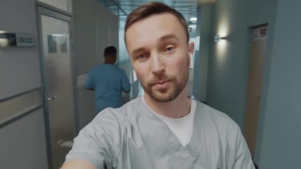 ПОВ молодого лікаря-чоловіка в медичній формі і бездротові навушники, що йдуть через коридор в лікарні, дивлячись на камеру і розмовляючи, маючи відеодзвінок або знімаючи відеоблог - Кадри, відео