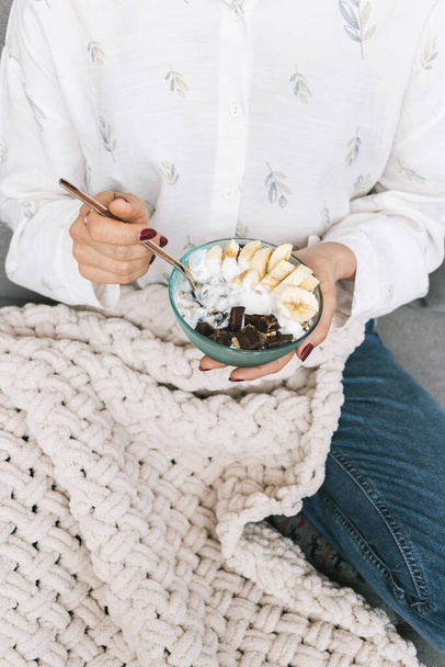 Mulher sentada em um sofá e comer alimentos saudáveis para o café da manhã, cereais, granola, muesli, aveia com banana, chocolate escuro e iogurte em uma tigela. - Foto, Imagem