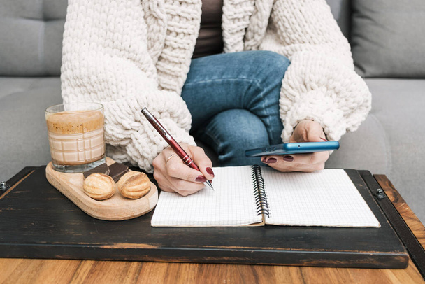 Femme méconnaissable assise sur un canapé à la maison, utilisant un téléphone portable ou un smartphone et écrivant dans un bloc-notes sur la table avec un verre de café et un dessert. - Photo, image