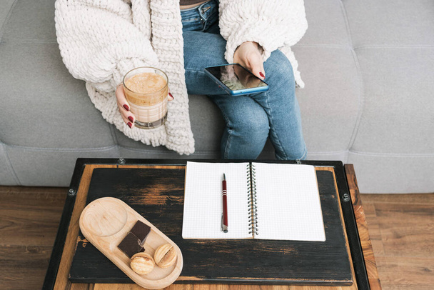 Femme méconnaissable assise sur un canapé à la maison, utilisant un téléphone portable ou un smartphone et tenant un verre de boisson au café. dessert et bloc-notes avec un stylo sur la table. - Photo, image