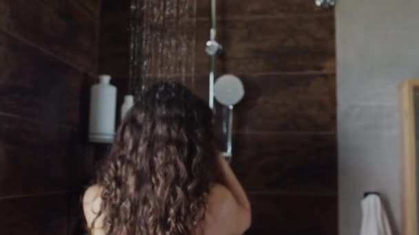 Foto trasera media de mujer joven irreconocible con el pelo largo y ondulado oscuro pisando bajo el agua que fluye en la ducha y remojando el cabello húmedo antes de lavarse - Metraje, vídeo