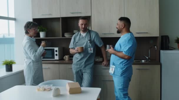 Група лікарів, які носять медичну уніформу, п'ють каву і спілкуються, відпочиваючи разом у кімнаті відпочинку під час робочого дня в лікарні - Кадри, відео