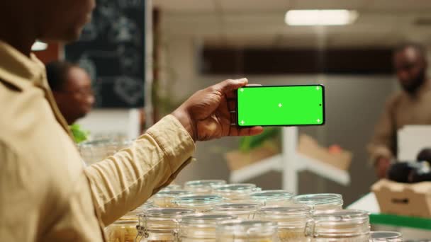 vendedor afroamericano muestra pantalla verde en el teléfono móvil en el mercado de barrio local, la venta de productos a granel naturales. El comerciante sostiene el dispositivo con la pantalla de maqueta del espacio de copia en blanco. Cámara A. - Metraje, vídeo