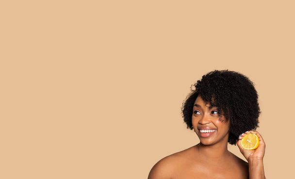 Zářící Afroameričanka drží čerstvý citrónový plátek vedle obličeje, radostně se usmívá, představuje přírodní výhody péče o pleť na béžovém pozadí, volný prostor, banner - Fotografie, Obrázek