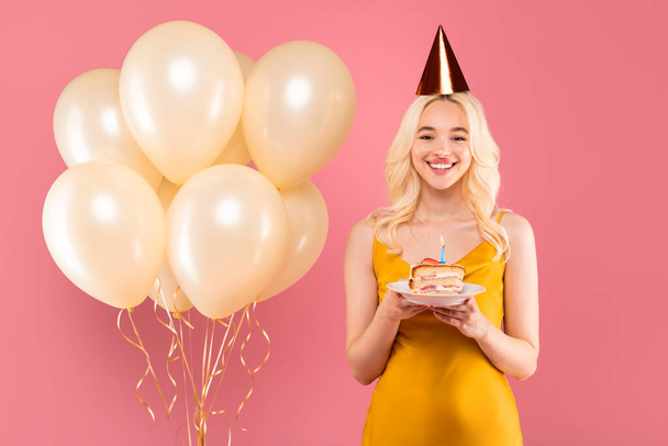 Mujer rubia radiante en vestido amarillo y sombrero festivo, sonriendo con plato de pastel al lado de globos dorados, celebrando en el fondo del estudio rosa - Foto, imagen