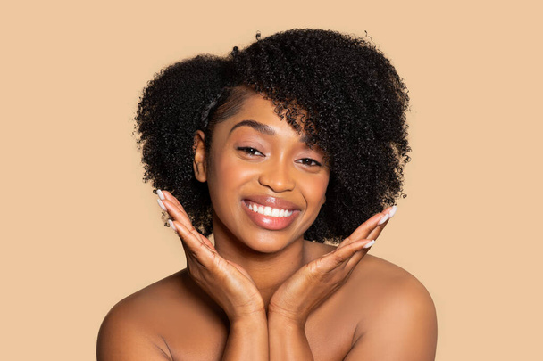 Stralende Afro-Amerikaanse vrouw met volumineus krullend haar brede glimlach, hand in hand nabij gezicht, poseren tegen beige studio achtergrond - Foto, afbeelding